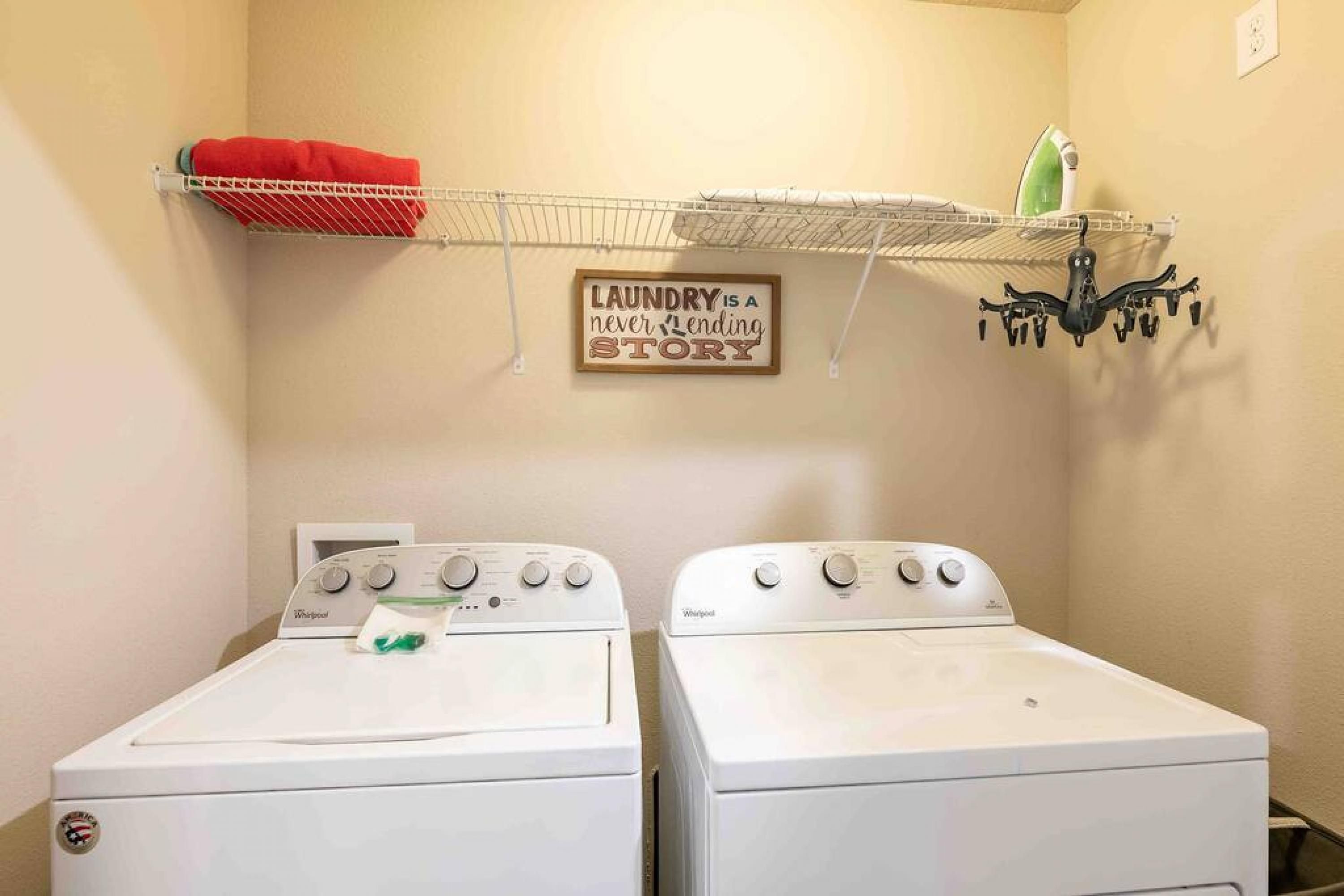 A laundry room with two Whirlpool washers, a red towel on a rack, a green iron, and a sign that says "Laundry is a never-ending story."