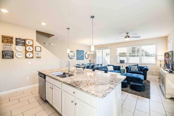 A modern kitchen with a granite countertop, white cabinets, and a built-in dishwasher. There is a dining area with a blue sofa and a city view through large windows. - Super Chic-4 Bedroom Townhome IN Disney Area (Kissimmee)