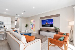 A modern living room with a sofa, armchair, and coffee table. There is a TV mounted on the wall and a kitchen area in the background.