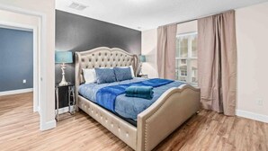 Une chambre à coucher avec un lit doté d’une tête de lit capitonnée, une table de chevet avec une lampe et une fenêtre avec des rideaux.
