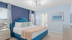 Un dormitorio con cabecera tapizada en azul, colcha color beige y mesita de noche blanca.