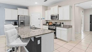 Una cocina moderna con armarios blancos, encimeras de granito, electrodomésticos de acero inoxidable y una isla central con taburetes de bar.