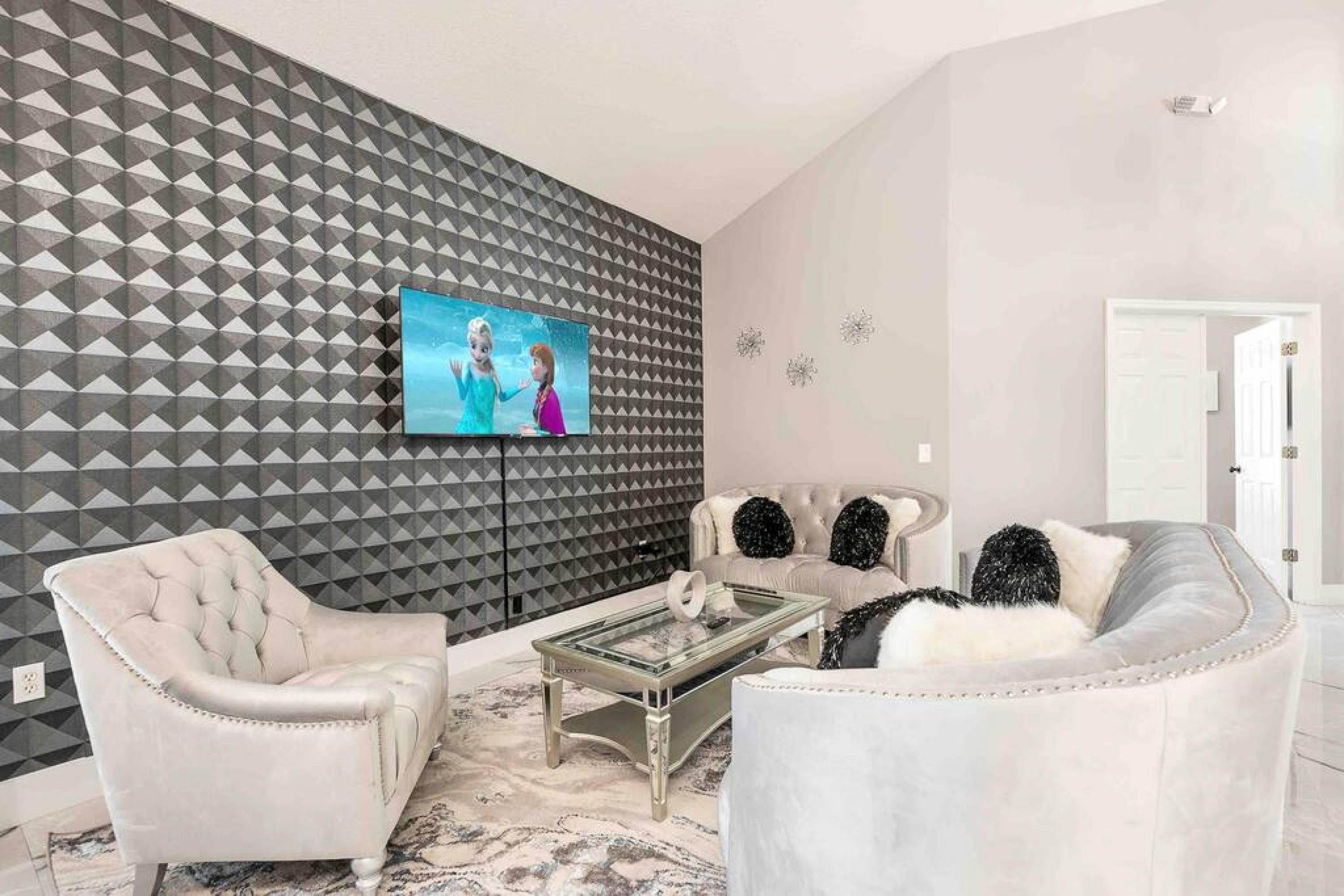 Een woonkamer met een geometrisch patroon op de muur, een flatscreen televisie, een gestoffeerde fauteuil, een bank met decoratieve kussens en een glazen salontafel.