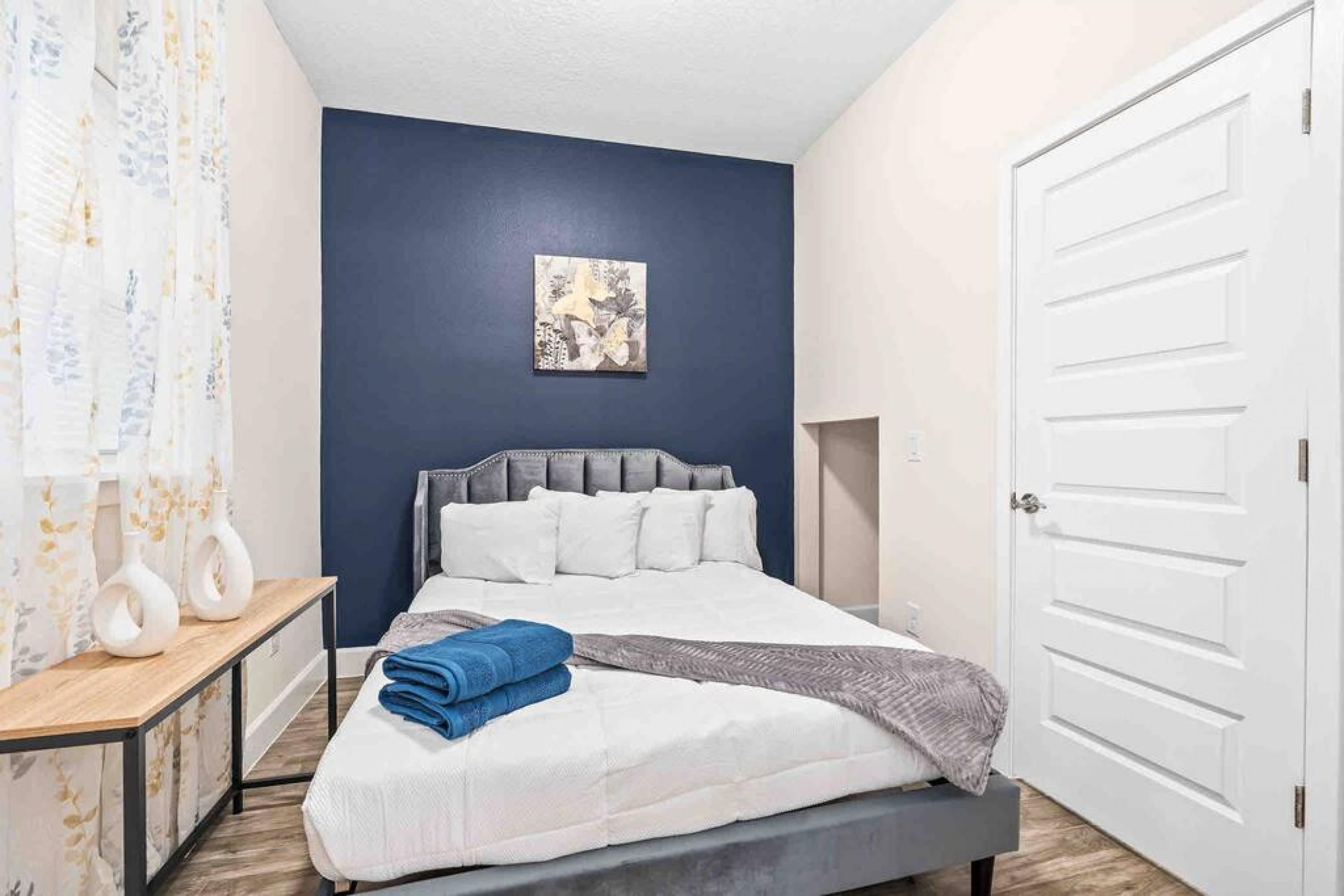 A bedroom with a bed, a bedside table, a nightstand, a wall-mounted artwork, and a door.