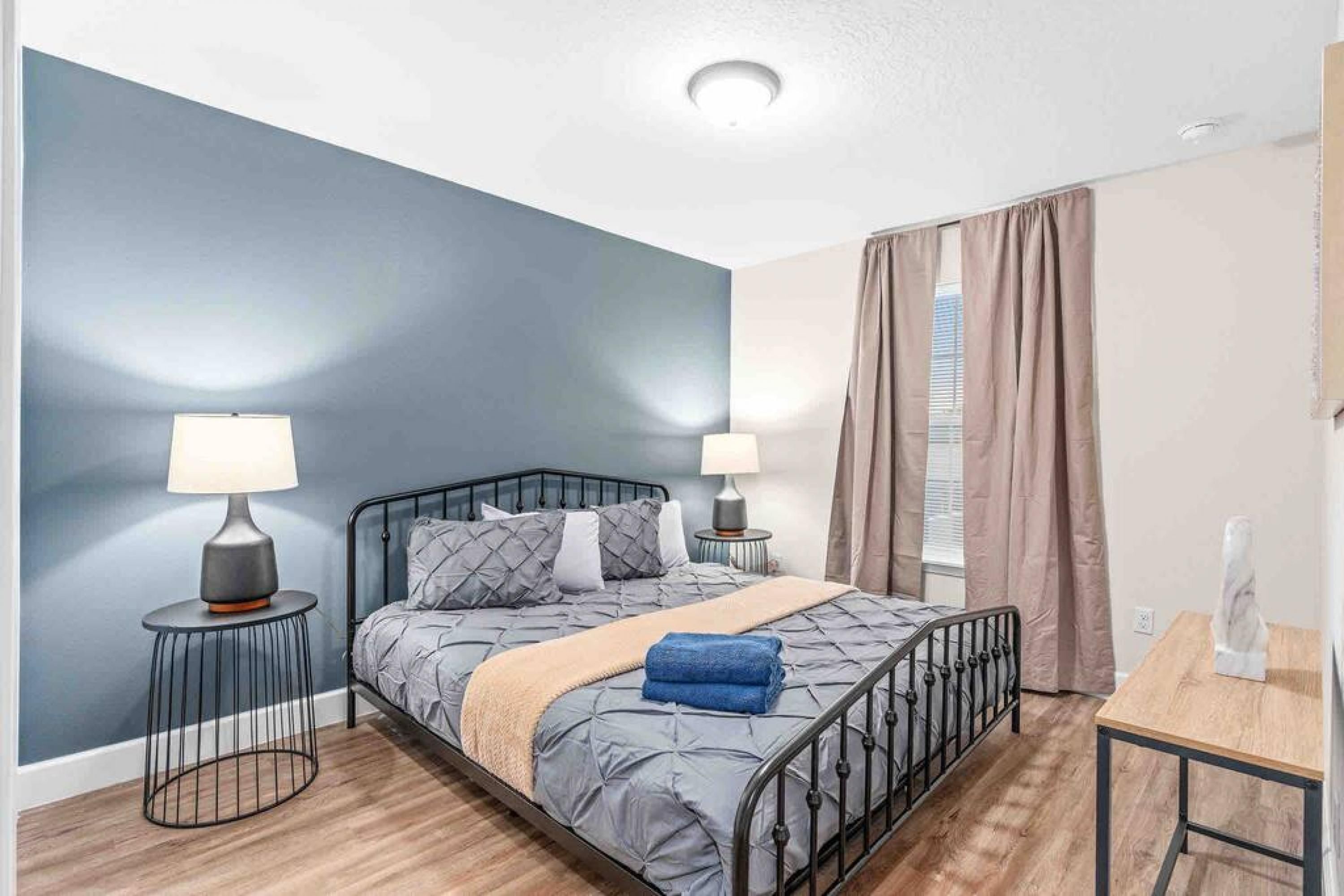 A bedroom with a bed, two bedside tables, a lamp, a window with curtains, and a wooden table.
