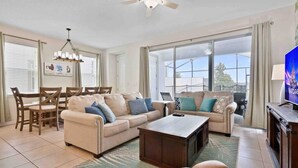 A spacious living room with a beige sofa, a coffee table, and a television. There is a dining area with a table and chairs, and large windows providing natural light. - Super Snug- FUN Themes Pool Tons OF Amenities (Kissimmee)