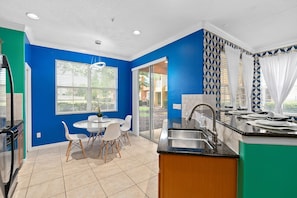 A modern kitchen with blue walls, a dining area with white chairs, and a large window with blinds. - Super Fly- Movie Loft w Recliners Near Disney (Kissimmee)