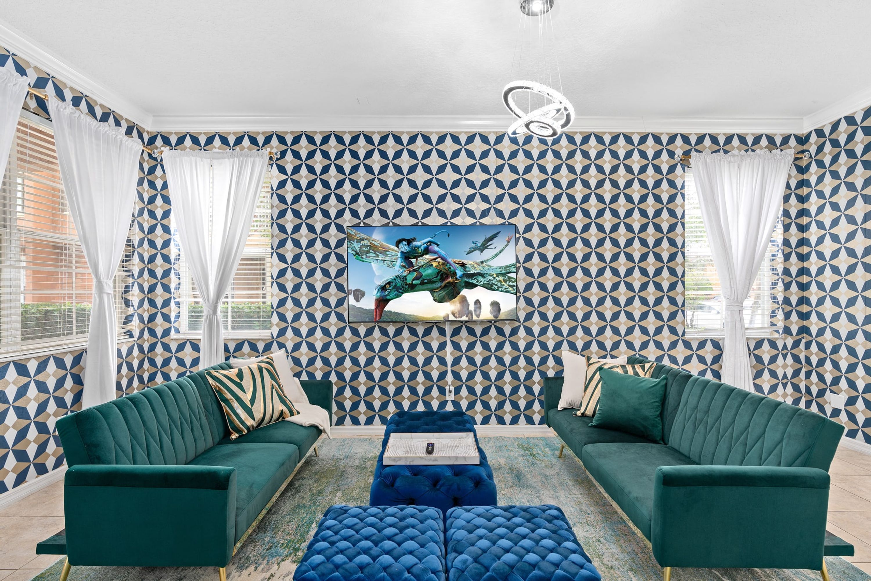 A room with a geometric patterned wallpaper, two green sofas, a blue ottoman, and a ceiling light fixture.