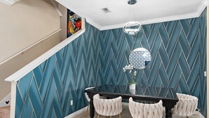 Un comedor con una mesa negra y cuatro sillas tapizadas con patas doradas. Las paredes tienen un papel tapiz azul turquesa con diseño de diamantes.