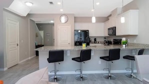 A modern kitchen with white cabinets, a stainless steel refrigerator, a microwave, and a coffee maker. There are three black bar stools and a large island with a granite countertop. - Super Liked - 4BD 3 5b 6 Miles To Disney Springs (Kissimmee)