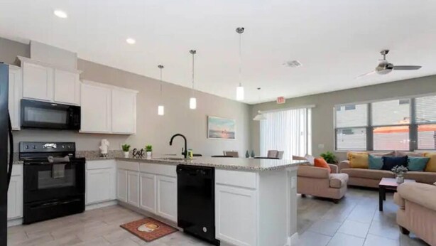 A modern kitchen with white cabinets, a black dishwasher, and a microwave. There is a dining area with a sofa and a coffee table. - Super Liked - 4BD 3 5b 6 Miles To Disney Springs (Kissimmee)