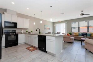 A modern kitchen with white cabinets, a black dishwasher, and a microwave. There is a dining area with a sofa and a coffee table. - Super Liked - 4BD 3 5b 6 Miles To Disney Springs (Kissimmee)