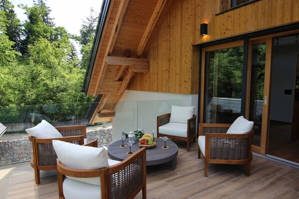 A wooden deck with wicker furniture and a round table, surrounded by glass doors leading to a covered area.
