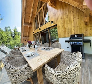 A wooden deck with a dining table set for four, surrounded by wicker chairs, and a barbecue grill.
