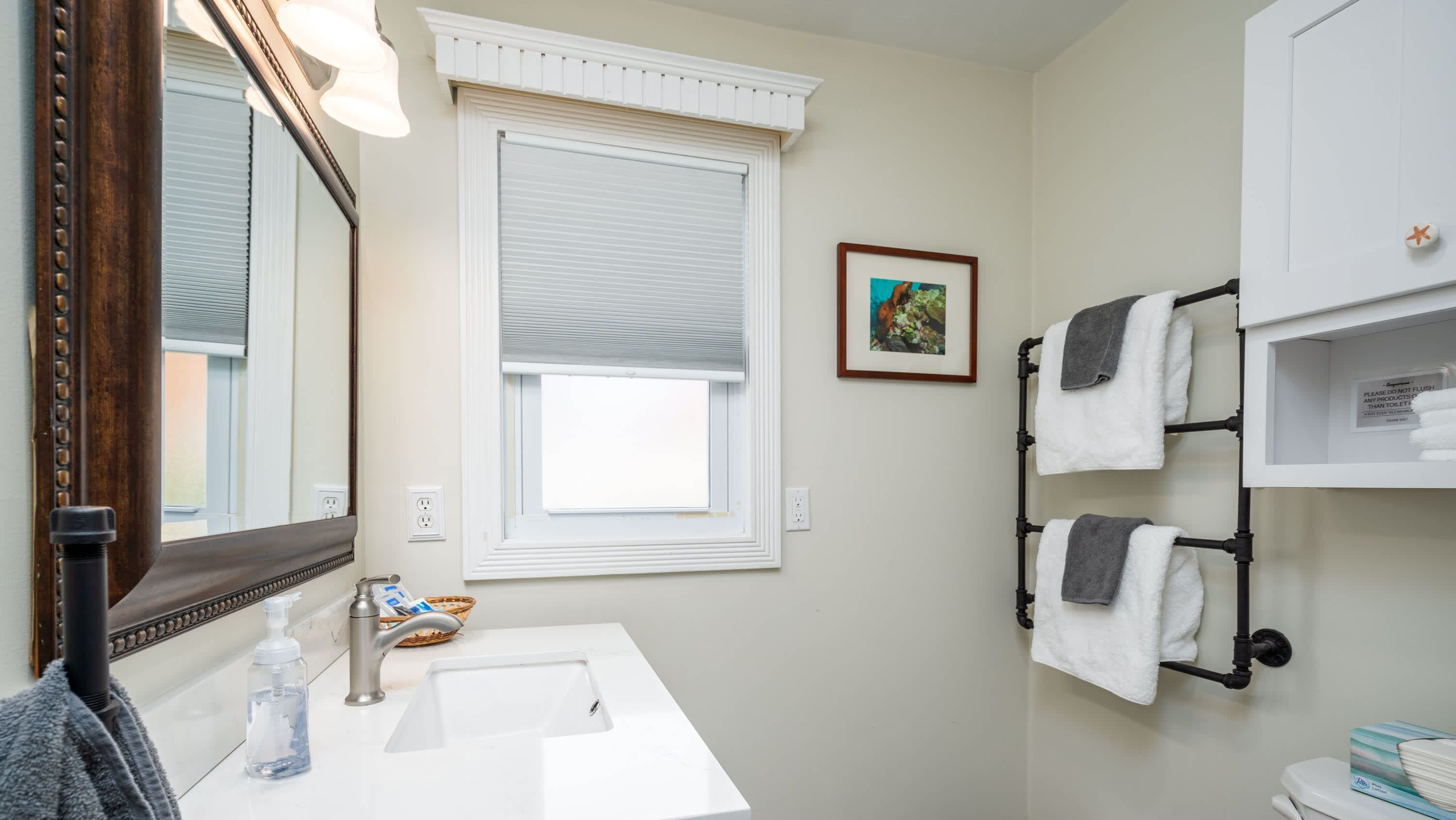 Un baño con un lavamanos blanco, un espejo, una ventana con persianas, un perchero para toallas y un cuadro en la pared.