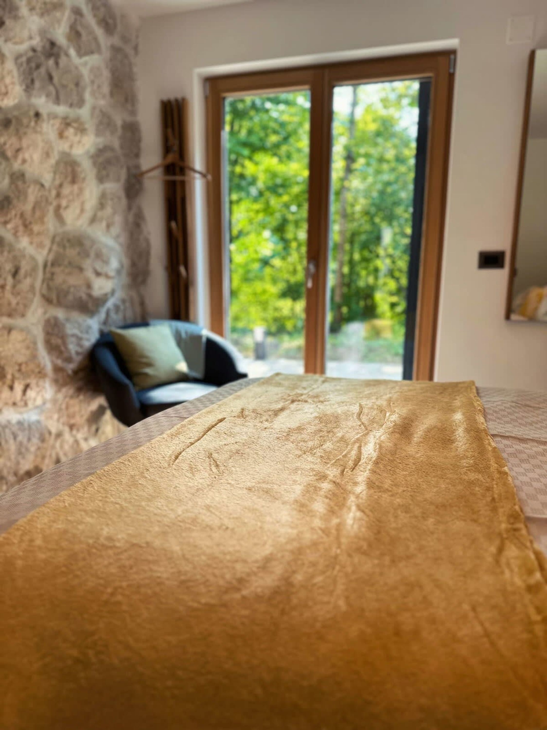 A room with a stone wall, a wooden door leading to a balcony with a view of greenery, and a bed with a patterned bedspread.