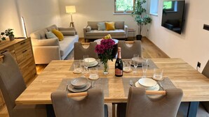 A dining area with a wooden table set for four, surrounded by chairs. There is a sofa set in the background, a TV mounted on the wall, and a window with natural light.