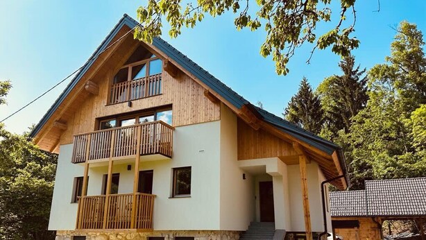 A two-story wooden house with a balcony, stone foundation, and a small garden area.