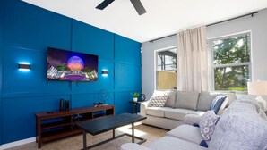 Ein modernes Wohnzimmer mit blauer Akzentwand, einem flachbildfernseher über einem Couchtisch aus Holz und einem grauen Sofa mit gemusterten Kissen.