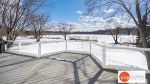 A wooden deck with a white railing overlooking a snowy landscape with trees and a clear sky. - Life is Better by the Water at Slayton Shores (Belding)
