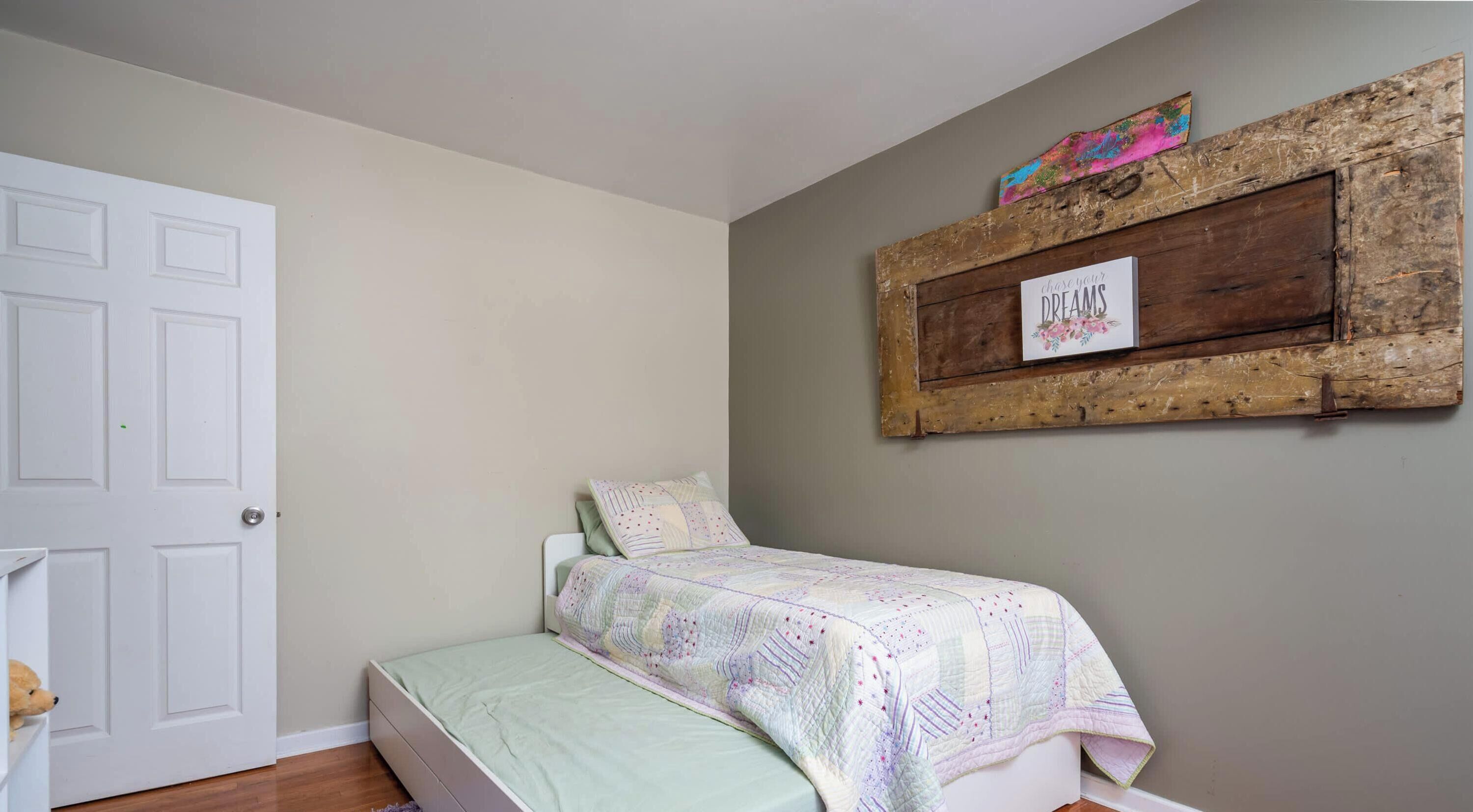 A bedroom with a bed, a white door, a purple rug, and a rustic wall decoration.