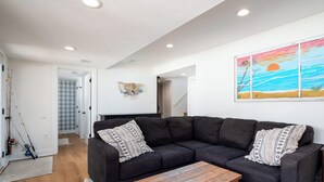 A modern living room with a dark sectional sofa, a wooden coffee table, and a painting of a sunset over the ocean. - Life is Better by the Water at Slayton Shores (Belding)