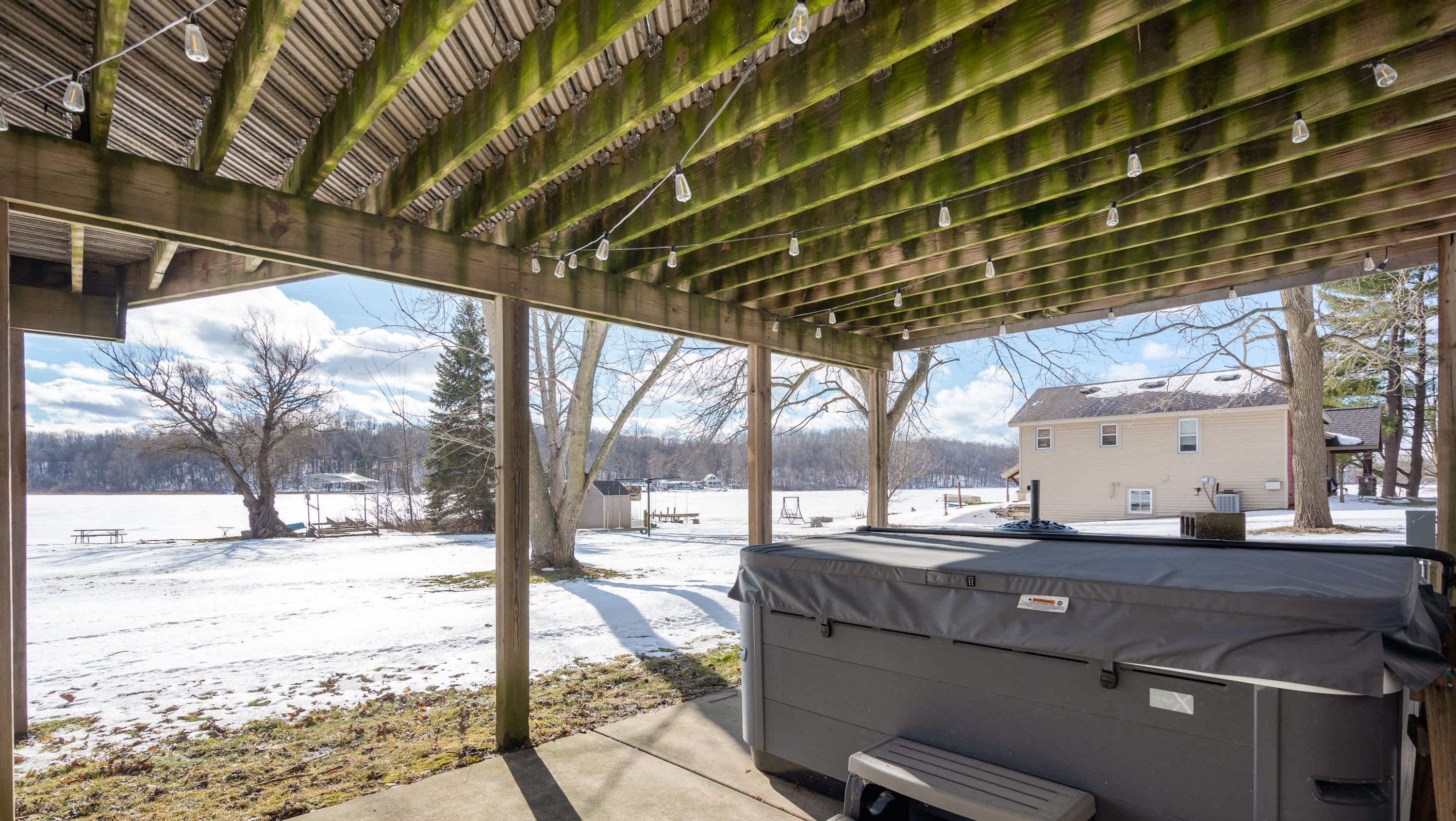 A covered outdoor area with a hot tub, benches, and a view of a snowy landscape and a house.