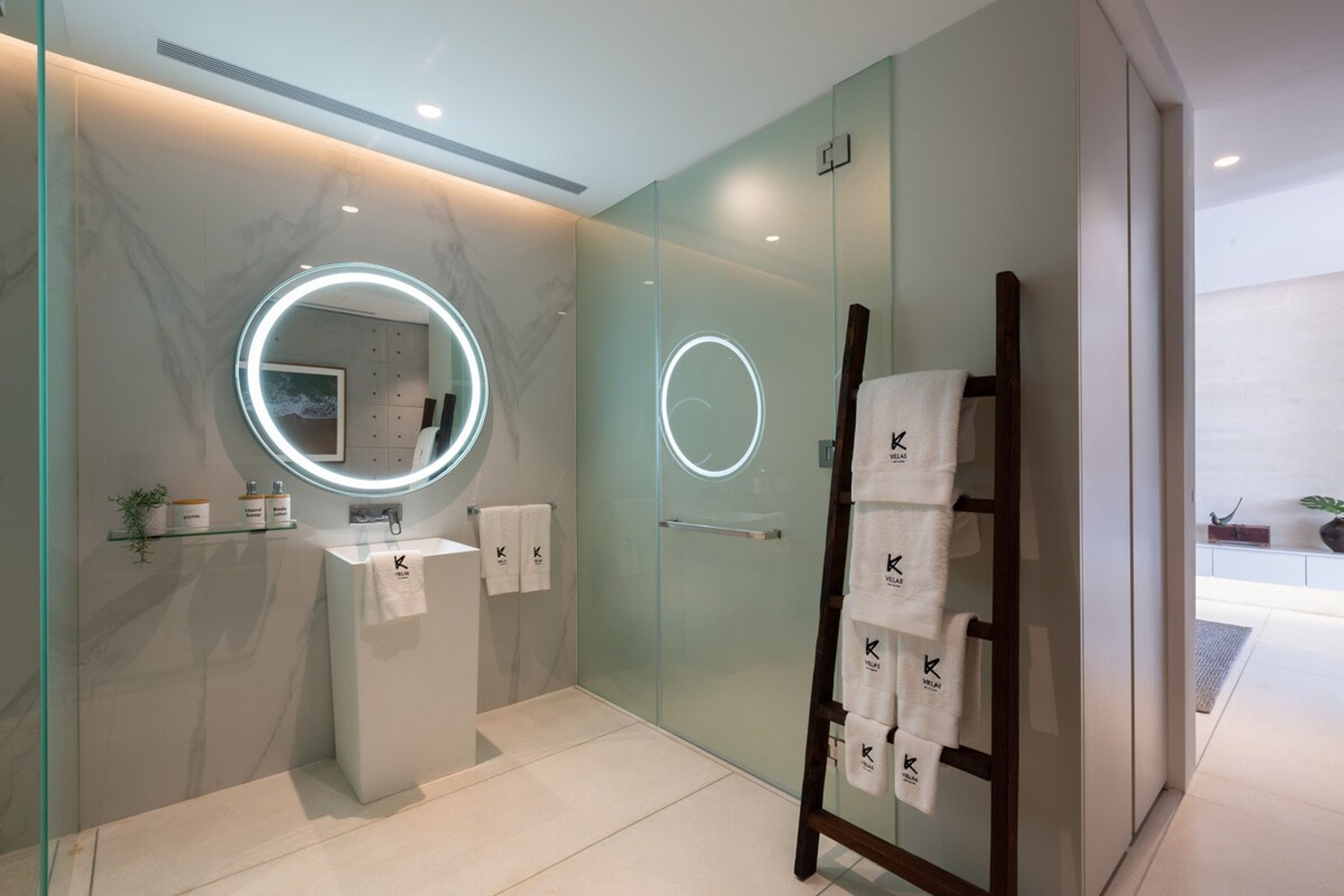 Un baño moderno con cabina de ducha de vidrio, un espejo redondo con iluminación y un perchero con toallas.