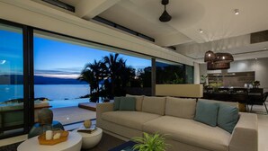 A modern living room with a large window overlooking a body of water, a sofa, a coffee table, and a dining area. - Syrah 3-bedroom Beachfront Villa in Koh Samui (Koh Samui)