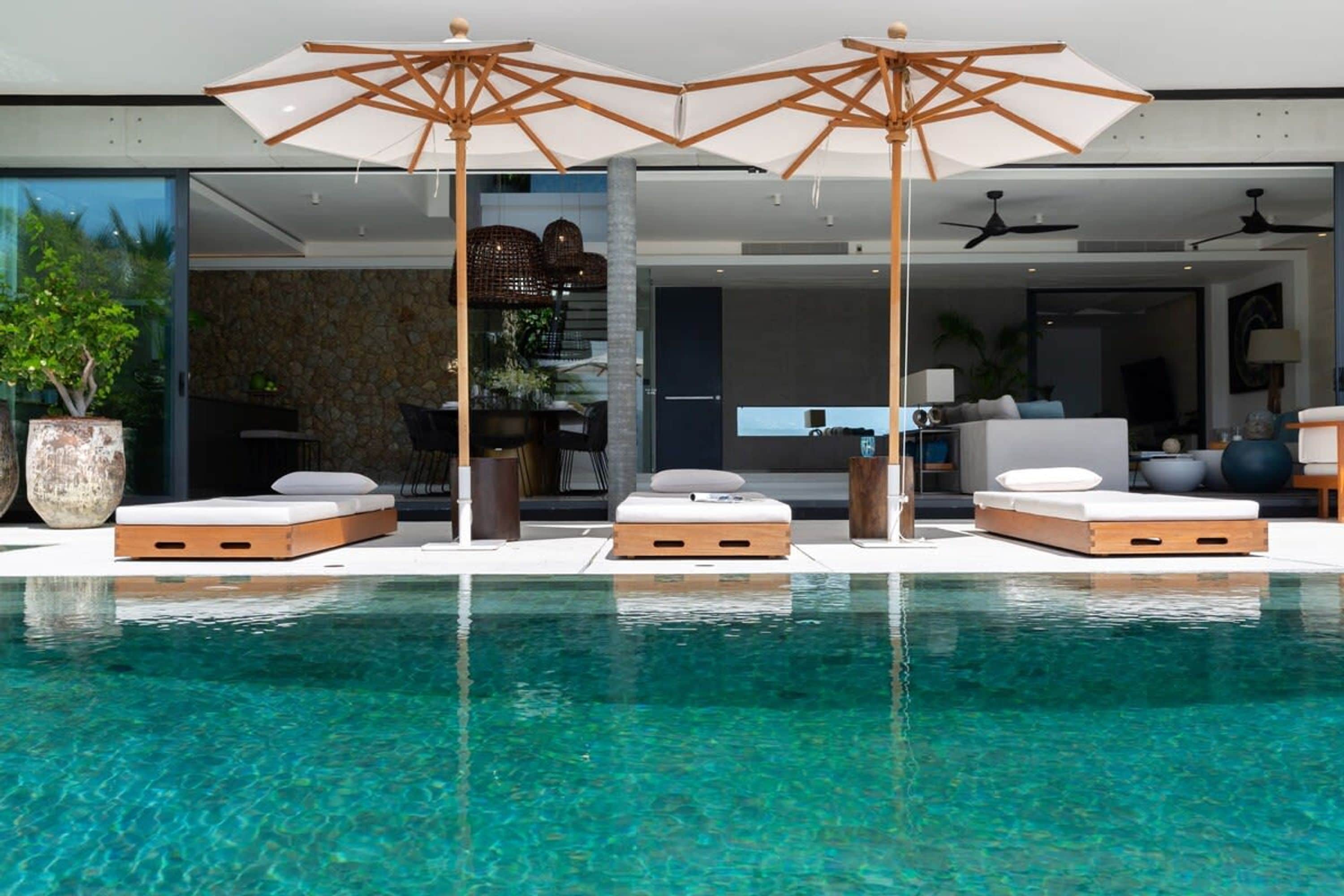 A modern pool area with clear water, three sun loungers, and two umbrellas.