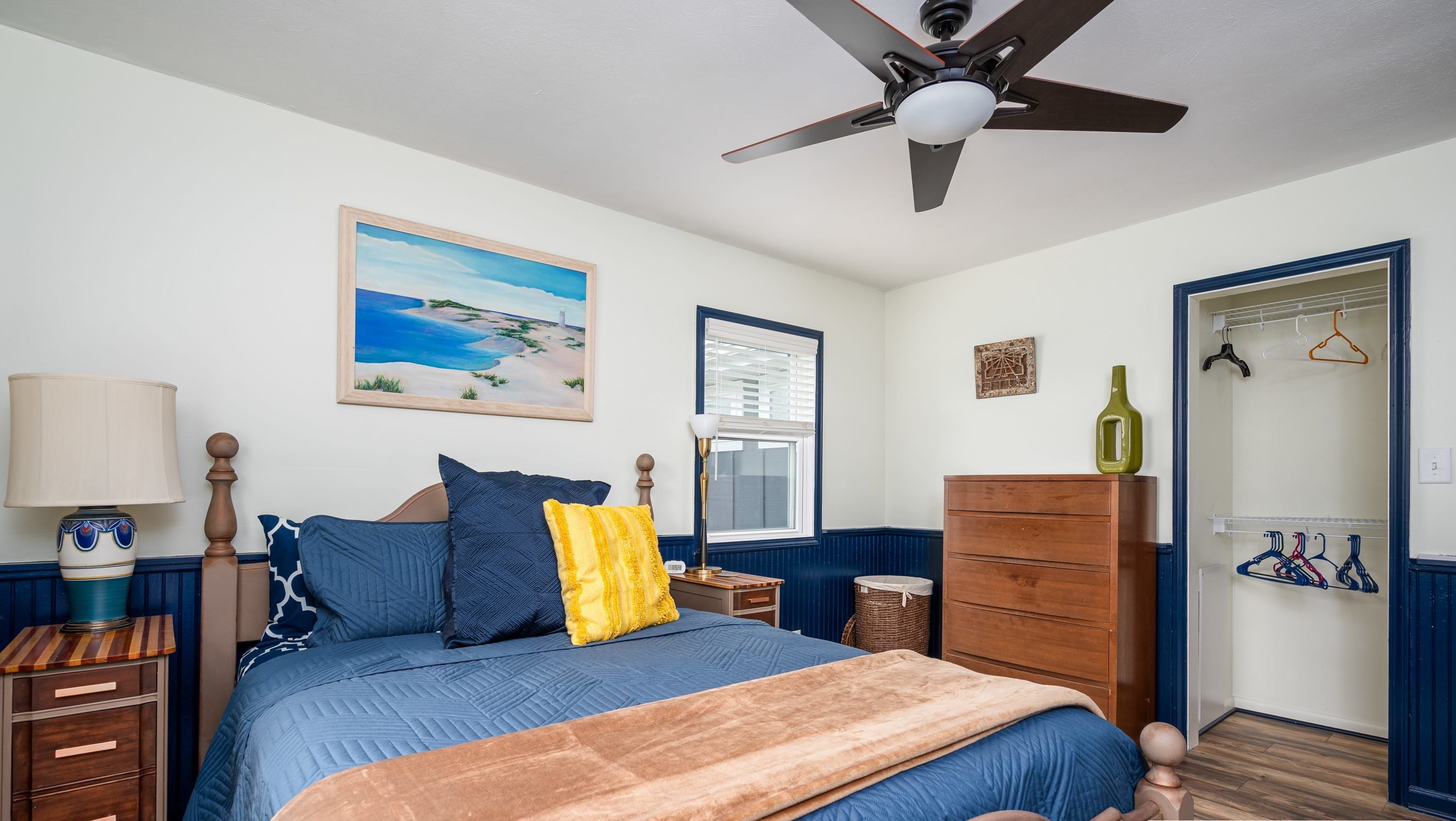 A bedroom with a bed, a ceiling fan, a dresser, a lamp, a window, and a painting.