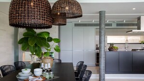 Un comedor moderno con una mesa de madera oscura, sillas negras y lámparas colgantes de mimbre. Hay una planta grande en maceta sobre la mesa, y se ve una cocina al fondo.