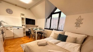 A bedroom with a bed, pillows, and a window. There is a kitchen area with a dishwasher, a television, and a dining table with chairs. - Charming City Loft Apartment With Free Parking (Ljubljana)