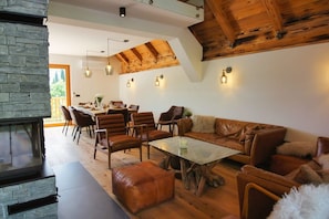 Un salon moderne doté d’une cheminée en pierre, de poutres apparentes en bois au plafond et d’un coin repas avec une grande table et des chaises.