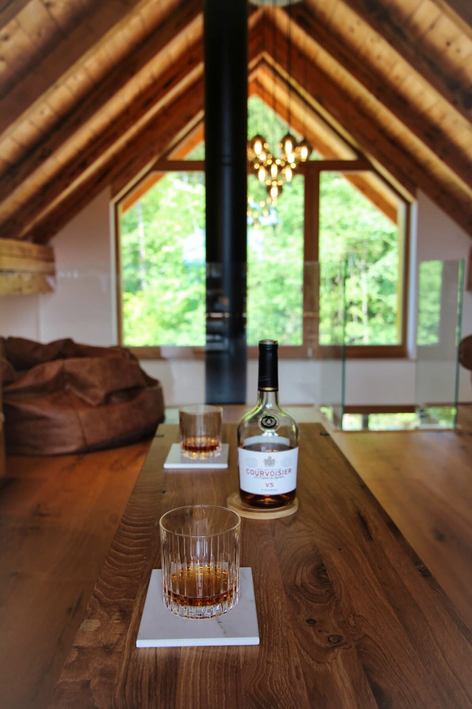 A wooden table with a bottle of liquor and two glasses, set in a room with a sloped ceiling and a view of greenery outside.