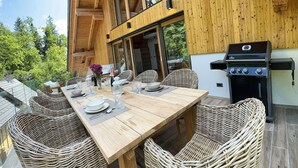 Eine Holzterrasse mit Esstisch für vier Personen, umgeben von Korbstühlen und einem Grill.