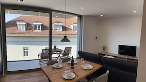 A modern living room with a dining area, large windows, and a view of a red-tiled building. - Garden Residence - Premium Downtown Apartment (Ljubljana)