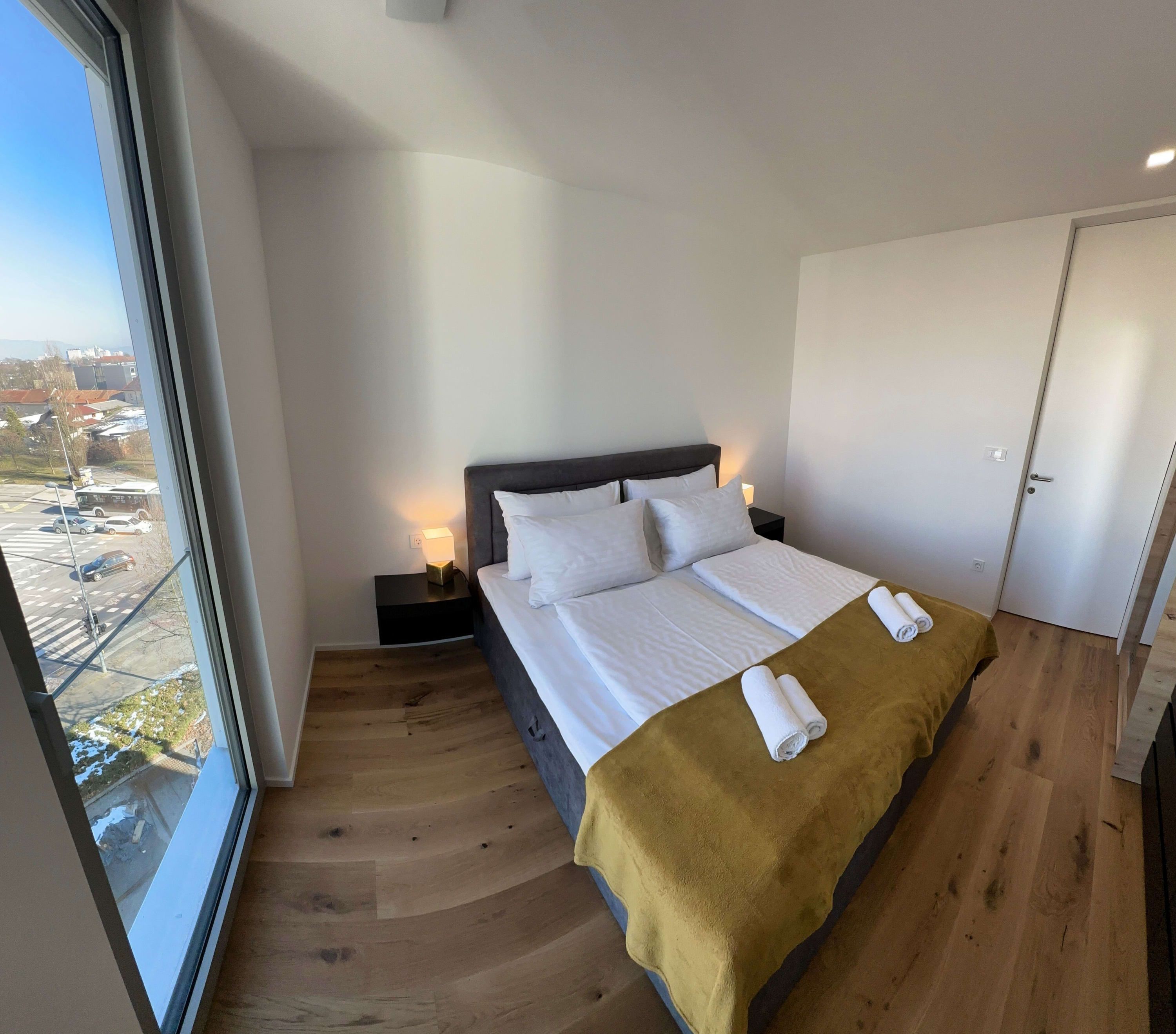 Une chambre d’hôtel moderne avec un grand lit, une table de chevet avec une lampe et une vue sur un parking et des immeubles, le tout visible à travers une grande fenêtre.