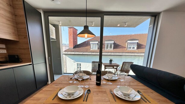 A dining area with a wooden table set for four, a black sofa, and a view of a residential building through a glass door. - Garden Residence - Premium Downtown Apartment (Ljubljana)