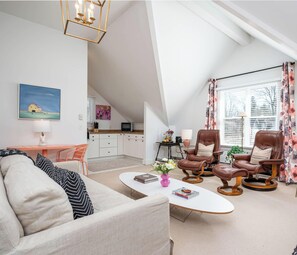 A modern living room with a high ceiling, a dining area, and a kitchen. There are two leather armchairs, a white sofa, and a coffee table. A painting and a lamp are on the wall.