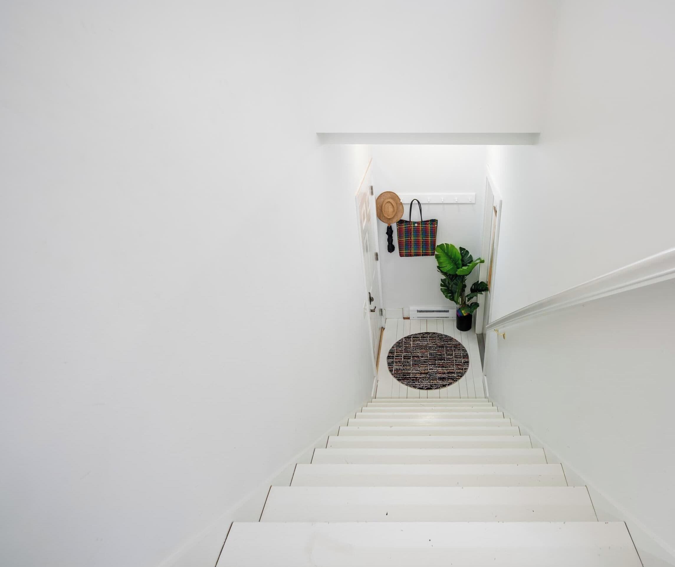 A white staircase leading to a landing with a door, a wall-mounted hat, a colorful woven bag, and a potted plant.