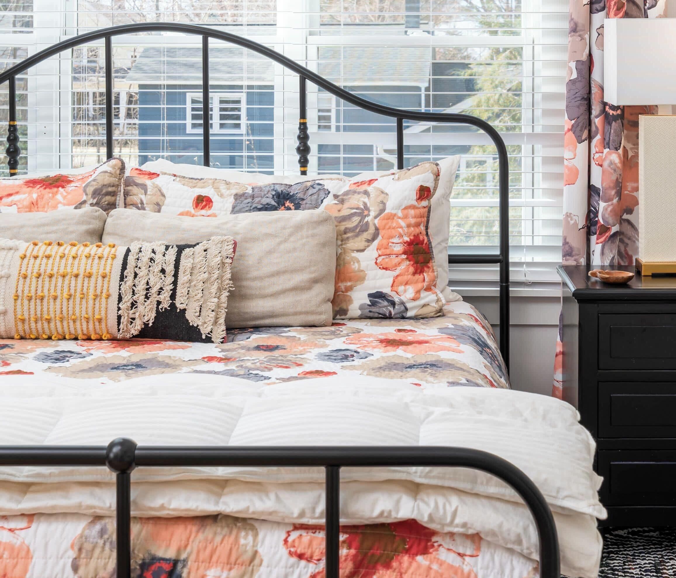 A bed with a floral-patterned bedspread, a beige throw pillow, and a yellow beaded throw.