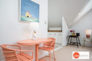 A dining area with a peach-colored table and chairs, a white wall with a painting, and a kitchen in the background.