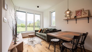 A modern living room with a wooden dining table, black chairs, a green sofa with cushions, a glass pendant light, and a large window with a view of greenery and mountains. - So a Valley Suites - Superior Apartment 09 (Bovec)