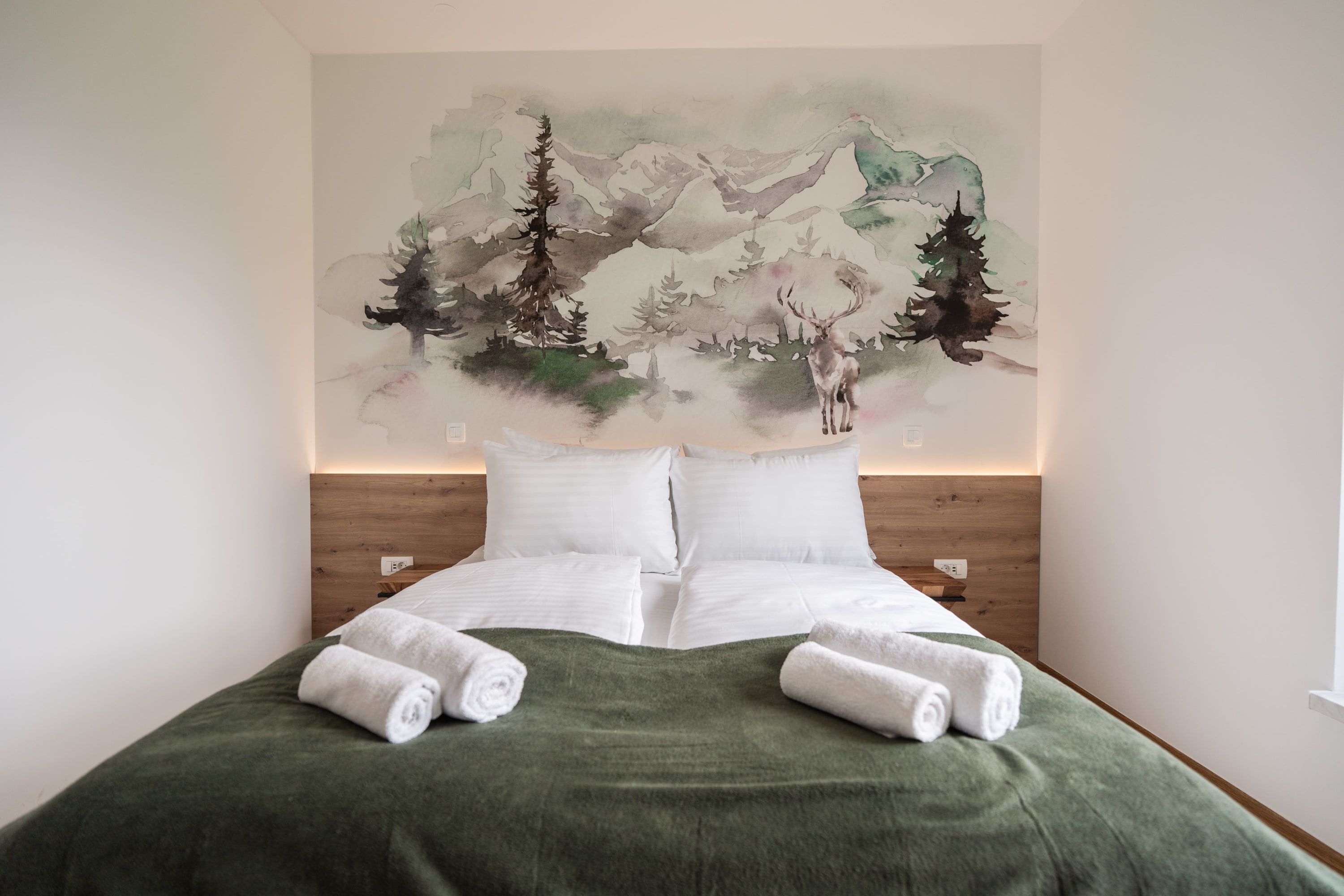 A neatly made bed with white linens and a green blanket, a large painting of a mountainous landscape on the wall behind, and a wooden headboard.