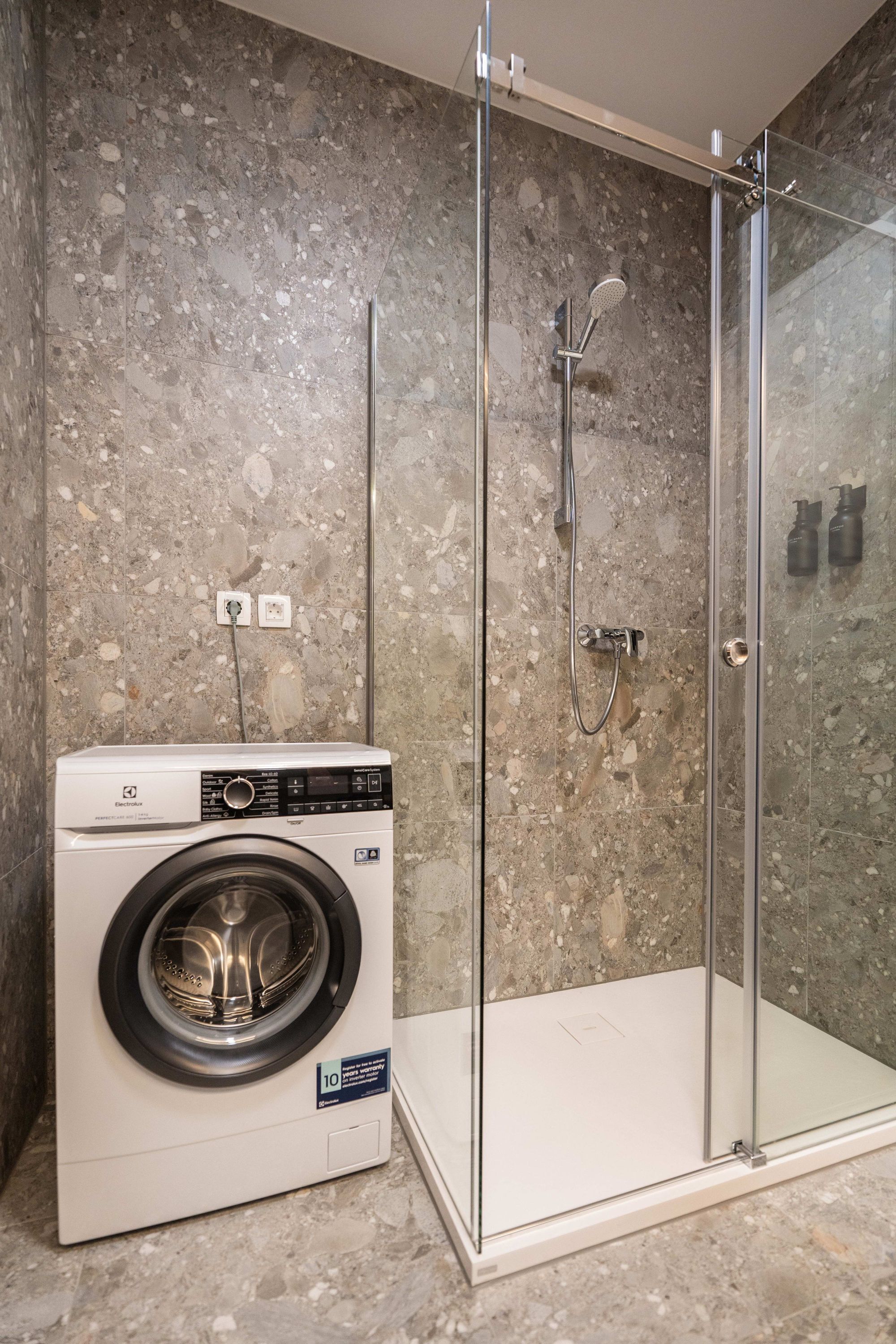 Un baño moderno con ducha de vidrio, lavadora integrada y pared de azulejos de mármol.