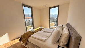Ein modernes Schlafzimmer mit Bett, großen Fenstern und Blick auf eine Stadtlandschaft.