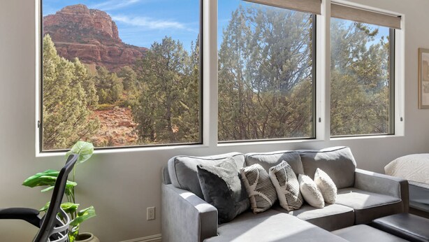 Un salón moderno con un sofá seccional gris, un sillón negro y una planta en maceta. Amplias ventanas ofrecen vistas a una formación rocosa roja y árboles.