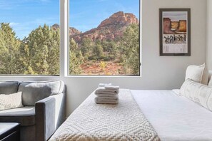 Una habitación de hotel con una cama, una silla, una ventana que ofrece vistas a formaciones rocosas rojas y árboles, y un cuadro en la pared.