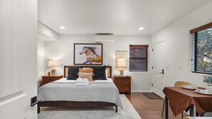 A bedroom with a bed, bedside tables, a window with blinds, a door, and a small dining area with a table and chairs. - Mammoth Rock Oasis-1 bd 1ba Hot Tub Free Pets (Sedona)